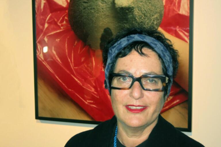 Middle-aged person with black-framed glasses and a blue headband, standing in front of a framed photograph featuring a greenish sphere on a red plastic sheet.