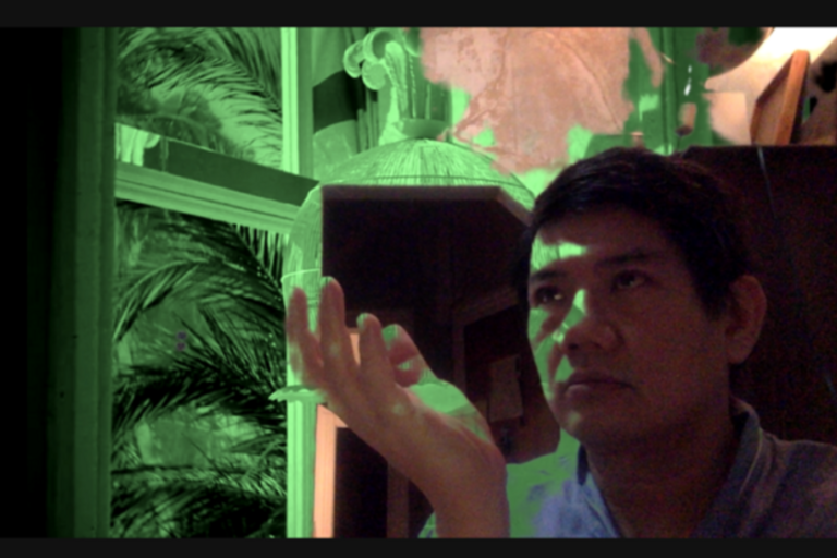 Man illuminated by green light, raising his open hand, with a window showing palm leaves in the background.