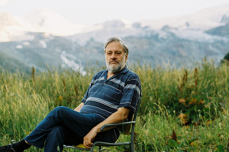 Older man with a grey beard seated outdoors in a meadow with a mountainous backdrop.