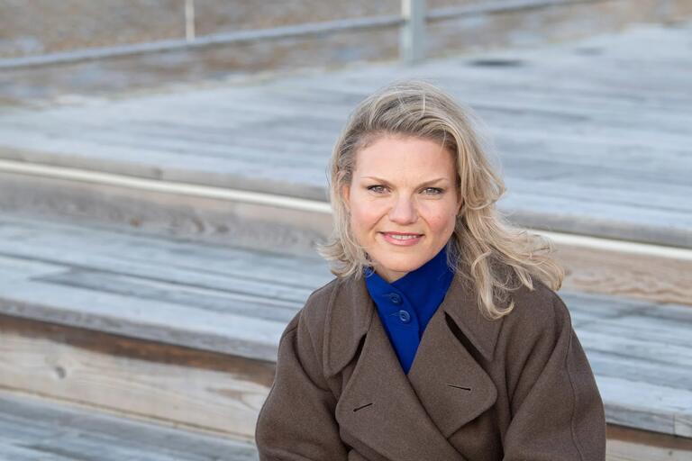 Woman with blonde hair wearing a brown coat and blue shirt, sitting on wooden steps outdoors.