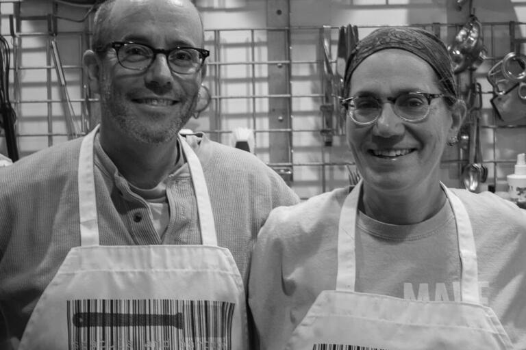 Two people smiling in a kitchen, wearing aprons with barcodes and spatula graphics.