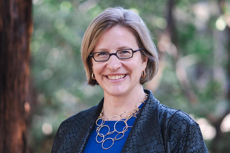 Woman with short light brown hair, wearing glasses, a dark textured jacket, and a geometric necklace, smiling outdoors with trees in the background.