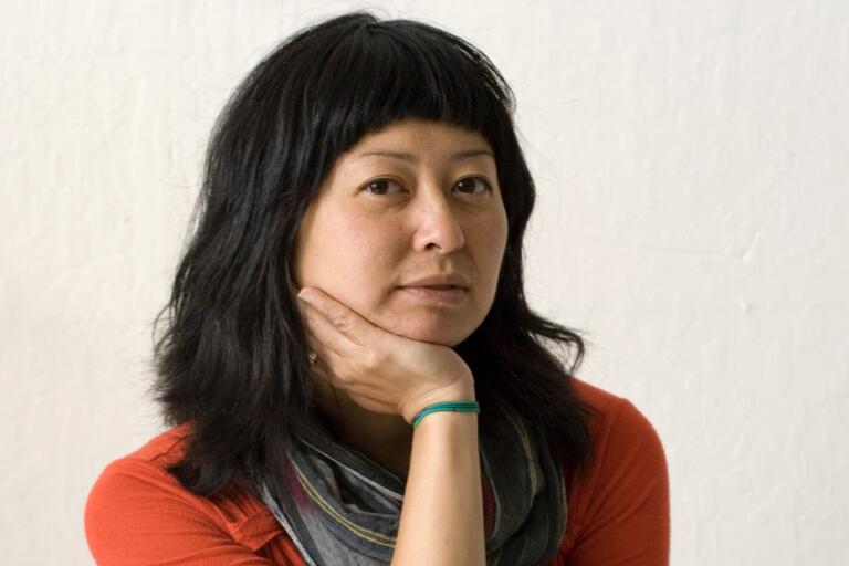 A person with black hair and bangs, wearing a red top and a multicolored scarf, rests their hand on their face against a plain, off-white wall.