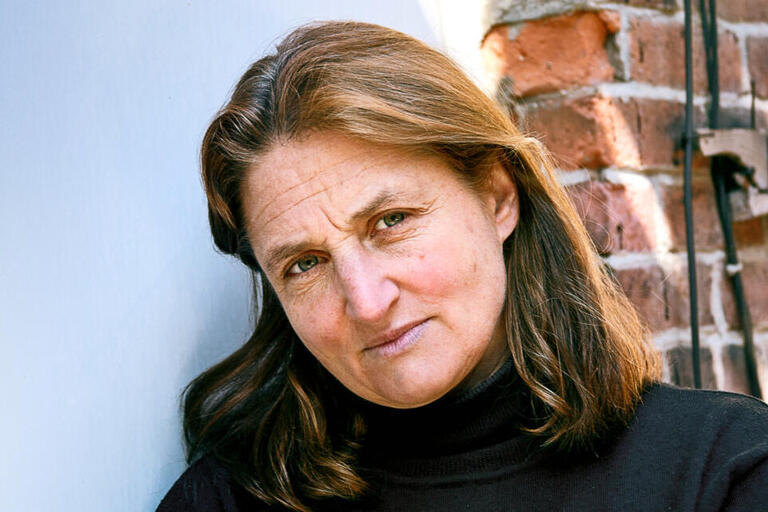 Middle-aged woman with long brown hair wearing a black turtleneck, standing next to a brick wall.