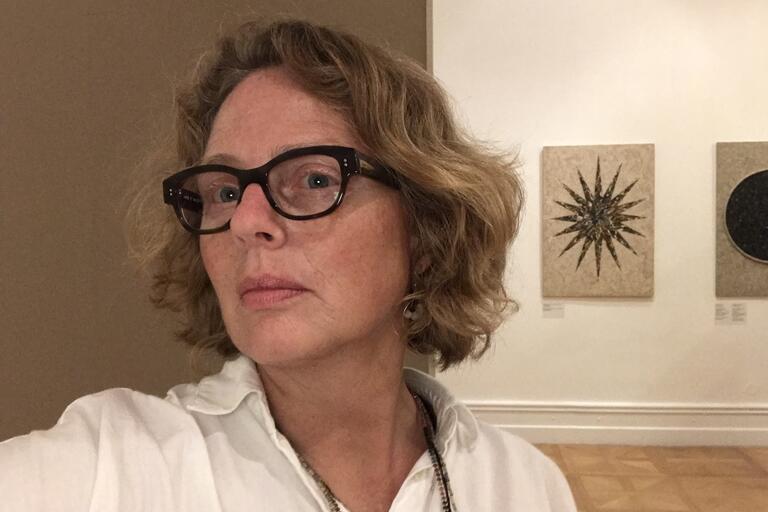 Close-up selfie of a person with glasses, short wavy hair, and a white shirt, in an art gallery with two artworks in the background.