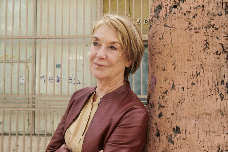 Older person in a brown leather jacket stands against a large tree trunk with barred windows and graffiti in the background.