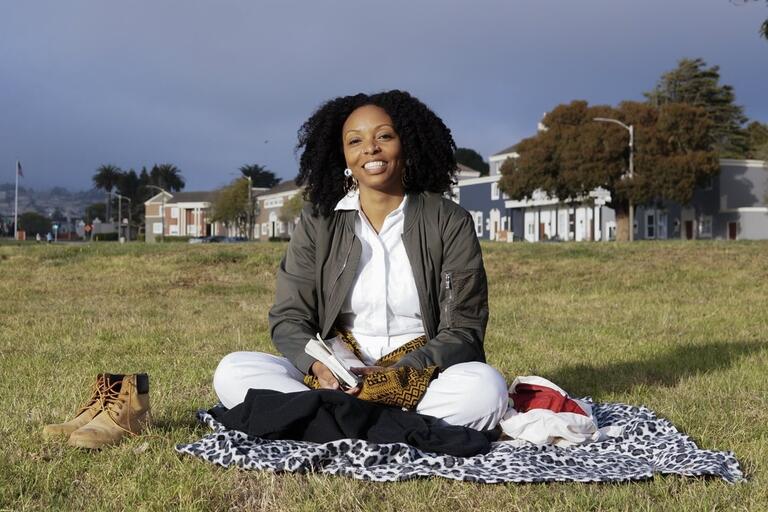 Woman sitting cross-legged on a blanket in a grassy field with an overcast sky.