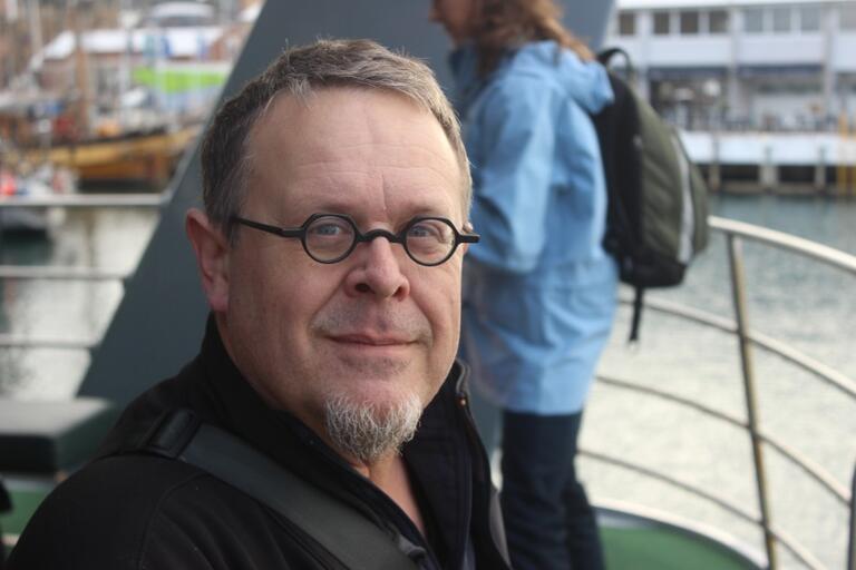 Man with glasses and a goatee on a boat deck, with another person and water in the background.