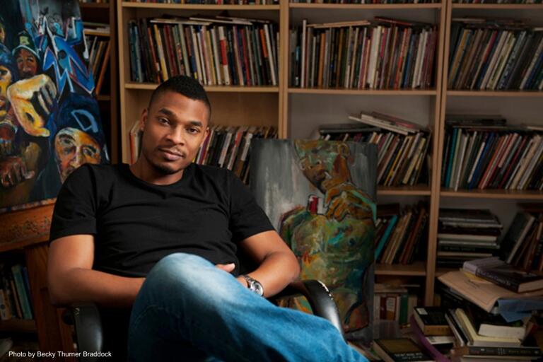 Man in black t-shirt and blue jeans sitting in front of bookshelves filled with books, with colorful artworks in the background.