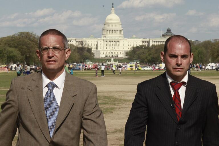 Two men in suits standing in front of the U.S. Capitol building on a sunny day.