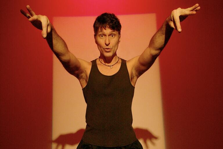 Person in a black sleeveless shirt with arms extended against a red and beige background.