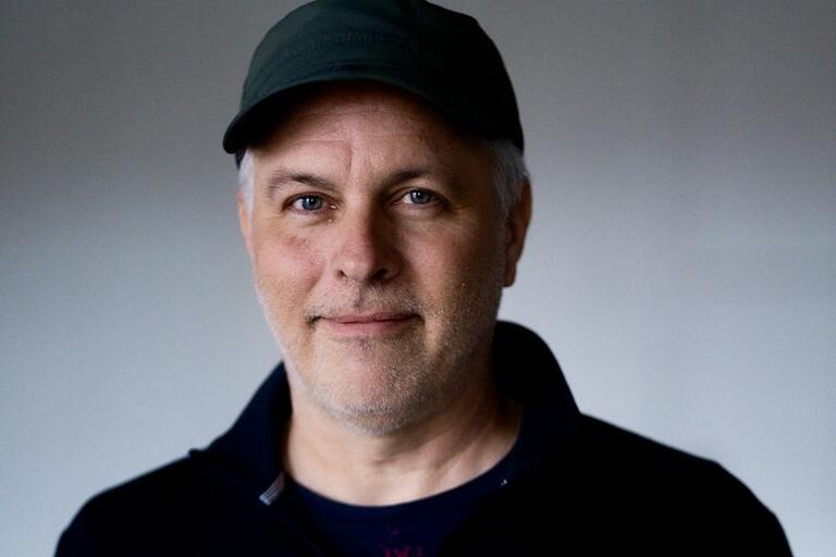Middle-aged man wearing a dark green cap and dark zip-up jacket against a plain background.