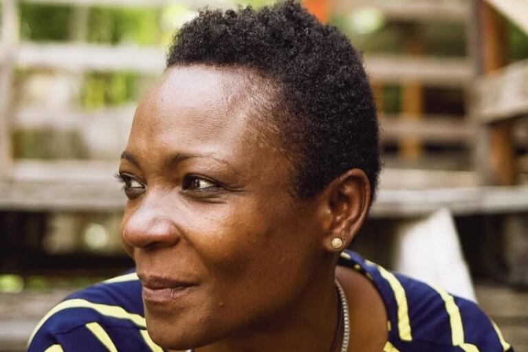 Close-up portrait of a person with short hair, wearing a navy blue shirt with yellow stripes, earrings, and a silver necklace, with a blurred outdoor background.