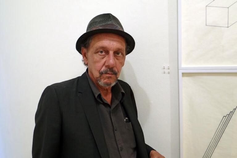 A man in a dark suit and fedora standing next to minimalist line drawings on a white wall.
