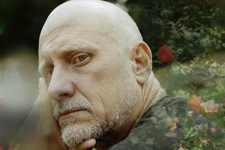 Close-up portrait of an older bald man with a gray beard, resting his face against his hand in a garden with blurred greenery and flowers in the background.