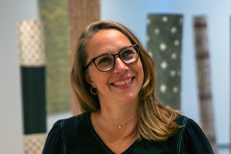 Smiling woman with glasses in a gallery-like setting with cylindrical patterned installations in the background.