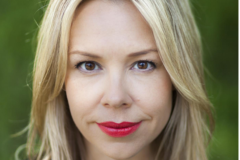 Close-up portrait of a person with light blonde hair, dark brown eyes, and red lipstick, against a blurred green background.
