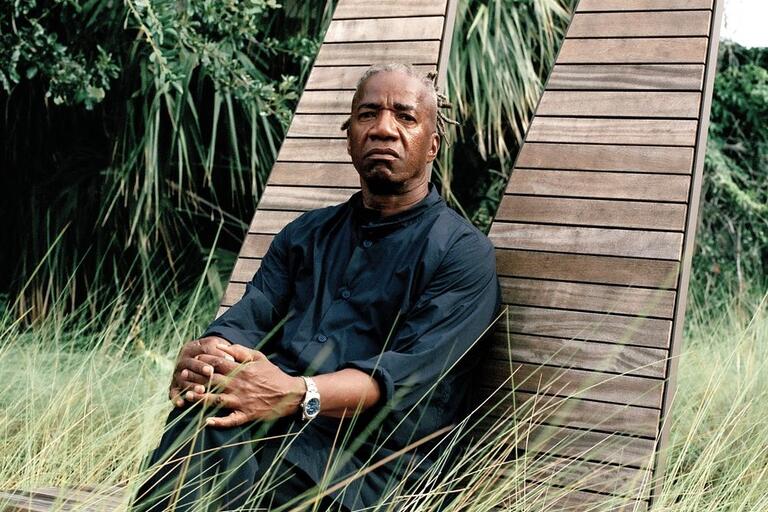 Man seated on a wooden outdoor chair in a lush, green setting with tall grass and trees.