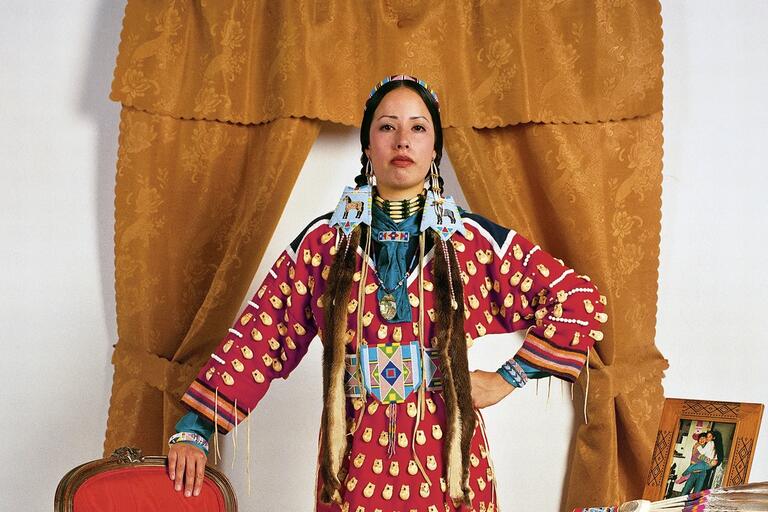 Person in traditional attire with a red outfit, beaded jewelry, and fur drapes, standing in front of golden-brown drapes.