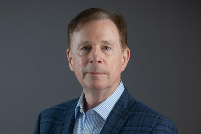 Headshot of a man with light brown hair wearing a blue plaid jacket over a striped dress shirt.
