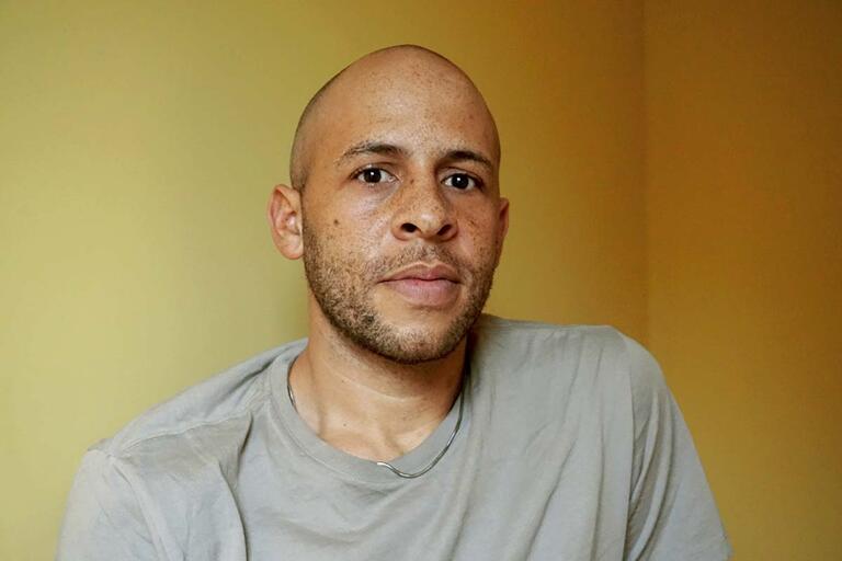 A man with a bald head and light beard wearing a gray t-shirt against a yellow wall.