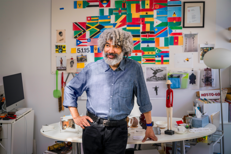 A man with gray curly hair and a beard stands in a colorful, cluttered office, smiling and resting his hand on a white desk. The background wall is covered with flags and various paper items.