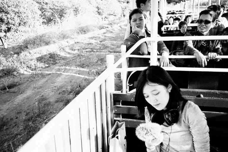Black-and-white photo of people on an outdoor train ride with the foreground focused on a woman holding a microphone.