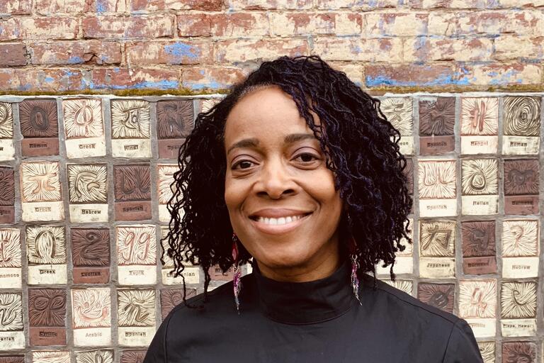 Woman smiling in front of a textured brick wall with labeled tiles showing various languages.