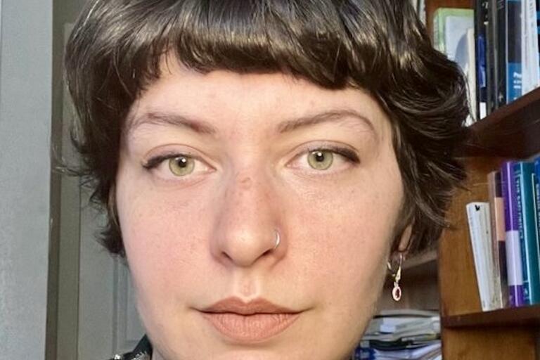 Close-up of a person with short brown hair, green eyes, a nose ring, and earrings, with a bookshelf in the background.