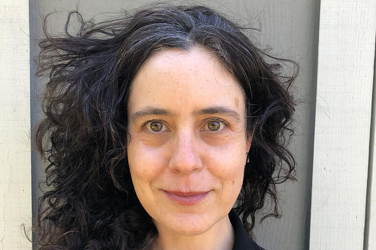 Close-up portrait of a person with dark, curly hair and a neutral expression against a background of wooden panels.