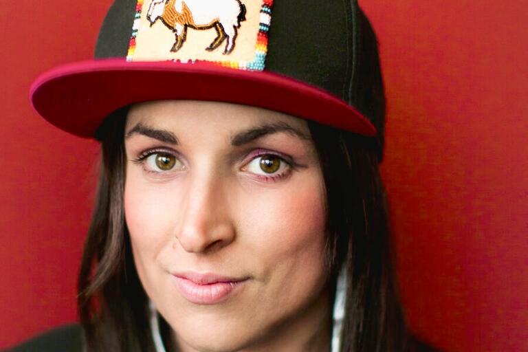 Close-up of a person with long dark hair wearing a black baseball cap with a beaded white buffalo patch.