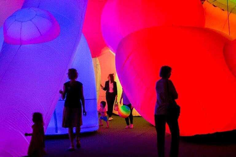 Silhouetted individuals explore a vibrant installation with large, illuminated, balloon-like structures in hues of purple, pink, and orange.