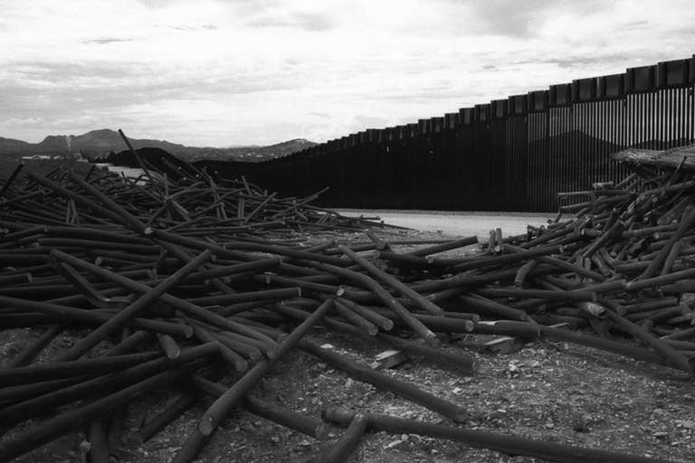 Black and white image of a pile of discarded metal poles in front of a tall border fence with mountains in the background.