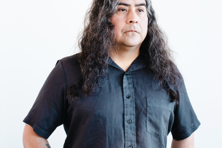 Raven Chacon has long, wavy hair wearing a black button-up shirt with a tattoo visible on their left arm, standing against a white background.