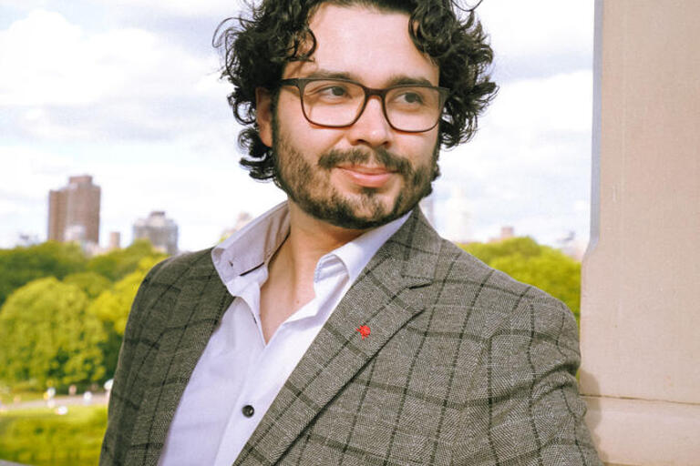 man in plaid jacket wearing glasses leaning against a wall with skyline behind him
