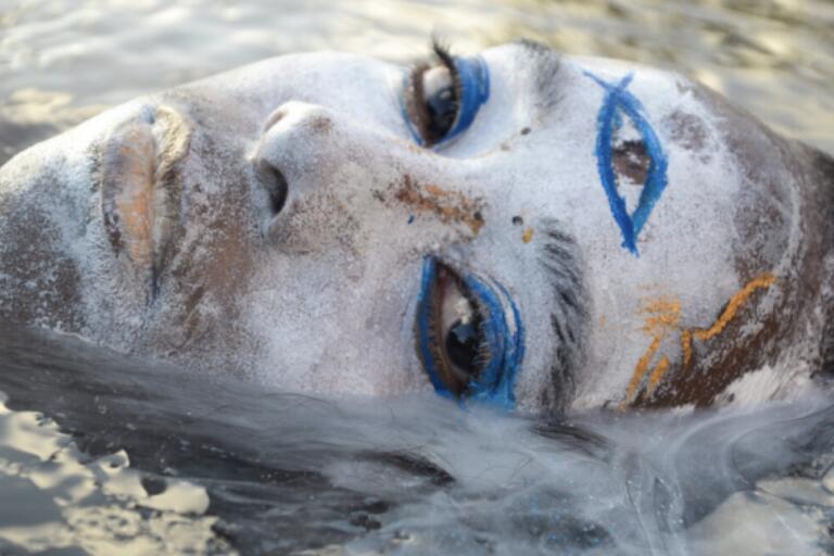 Close-up of a person's face submerged in water with dramatic blue eye makeup, silver face paint, and painted symbols on their forehead.