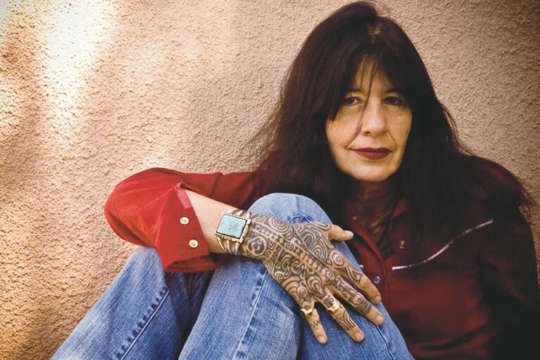 A person with long dark hair, wearing a red shirt and jeans, showcases a tattooed hand with a turquoise bracelet.