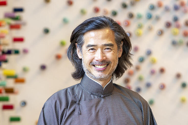 A man with long hair wearing a black shirt smiles warmly at the camera, exuding a friendly and approachable demeanor.