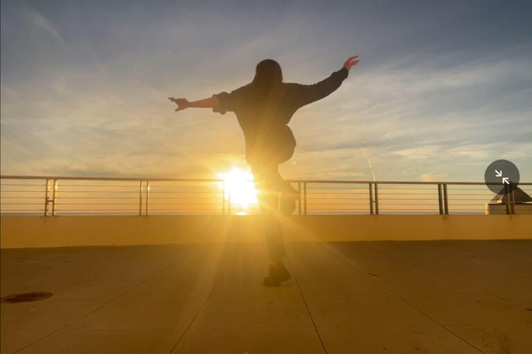 A silhouette of a jumping with arms outstretched, facing the sun as it sets on the horizon. The sun's rays are shining through the railing, casting long shadows on the ground.