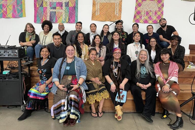 A diverse group of people in various traditional and contemporary outfits seated in three rows with colorful quilted artwork displayed on the wall behind them. Several individuals wear intricate necklaces or pendants, and traditional patterned garments.