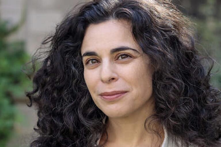 A woman with curly hair wearing a white jacket, standing confidently with a neutral background.