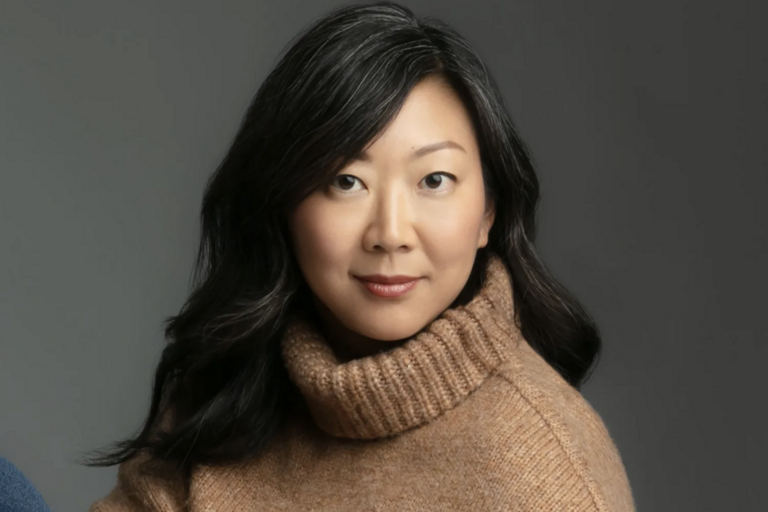 Asian woman wearing a cozy sweater and jeans, standing confidently with a warm smile.