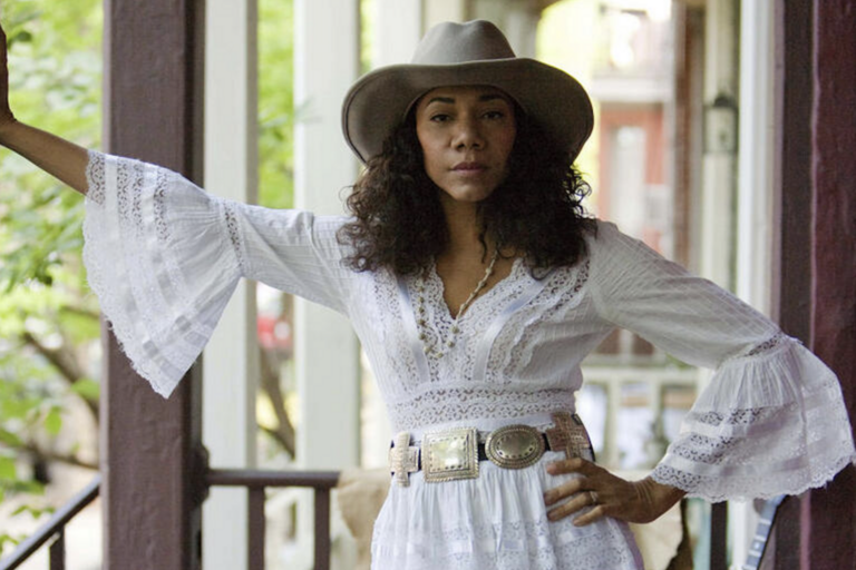  Person in a white lace dress and hat standing on a porch.
