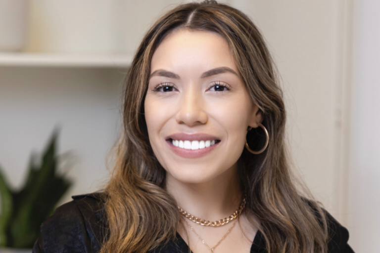 Smiling woman with long brown hair wearing hoop earrings and layered necklaces.