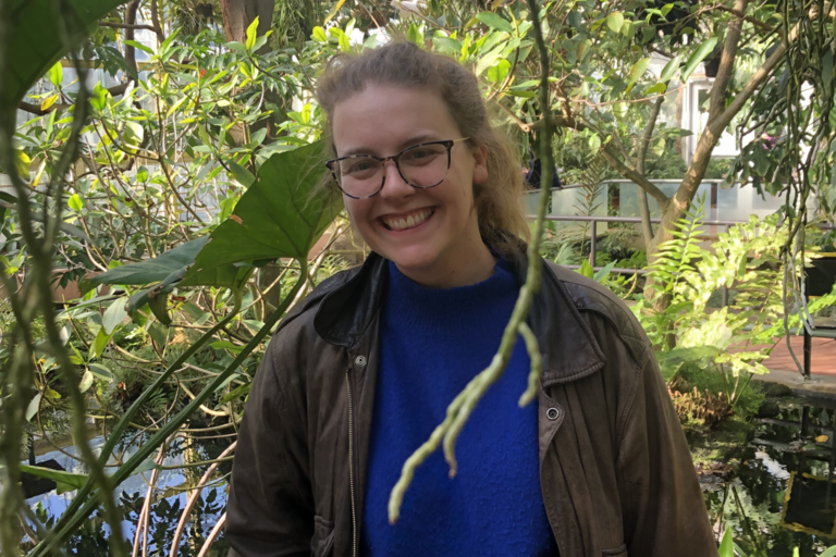 Smiling person with glasses and a blue sweater stands in a lush greenhouse filled with vibrant green plants. The atmosphere is serene and joyful.