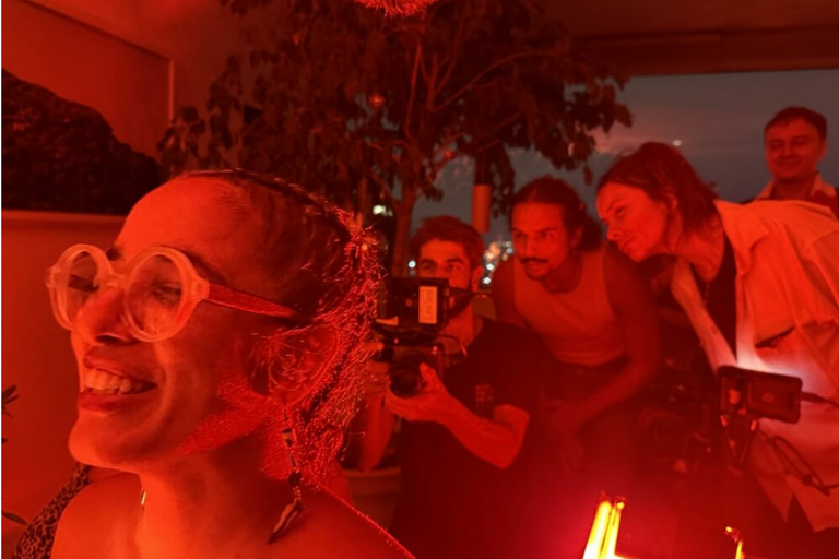 Person smiling with large glasses in a red-lit room, three people in the background focusing on a camera.