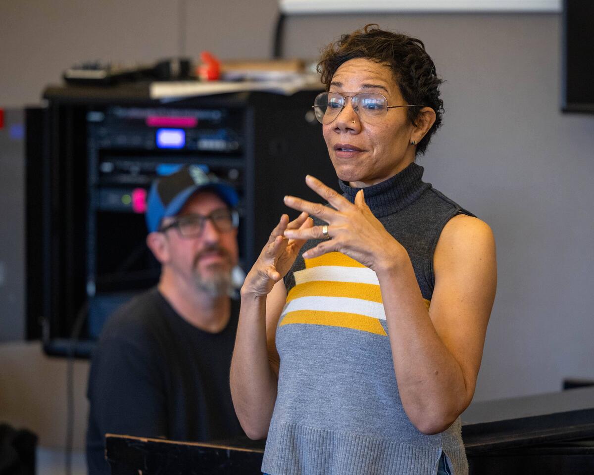 A woman gesturing while speaking, with a man in the background.