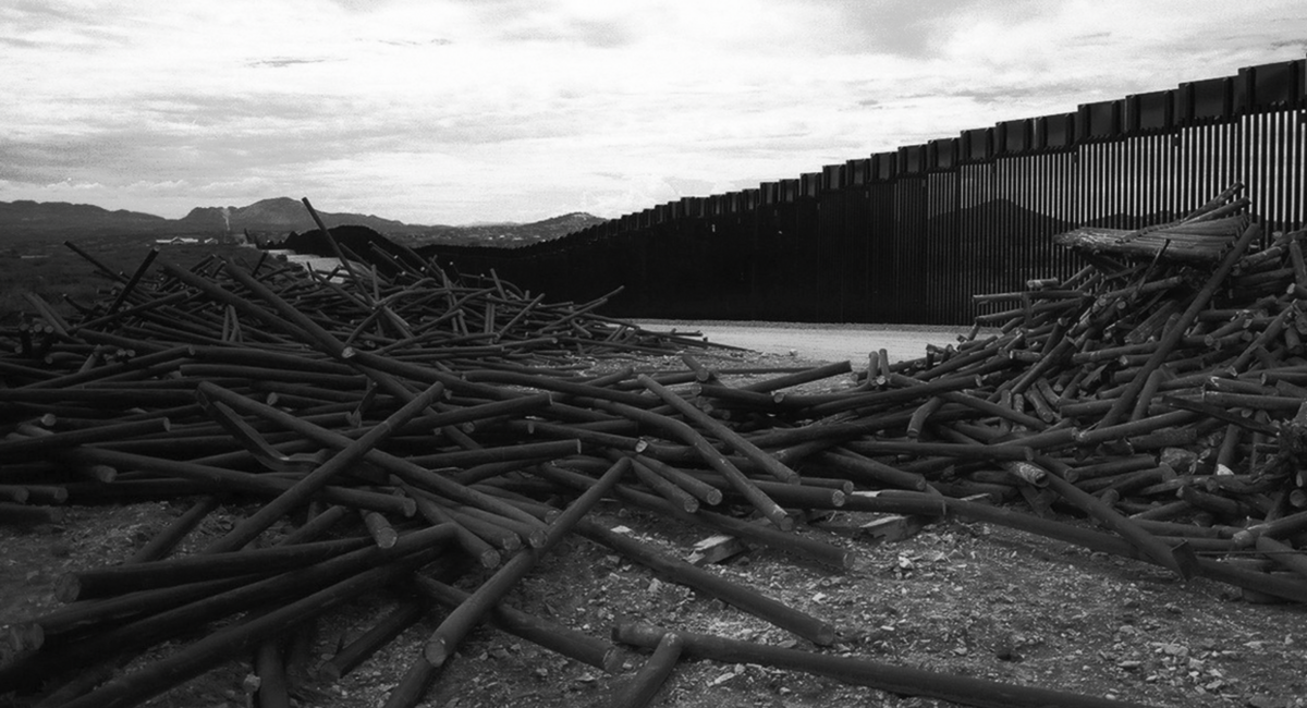 Black and white image of a pile of discarded metal poles in front of a tall border fence with mountains in the background.