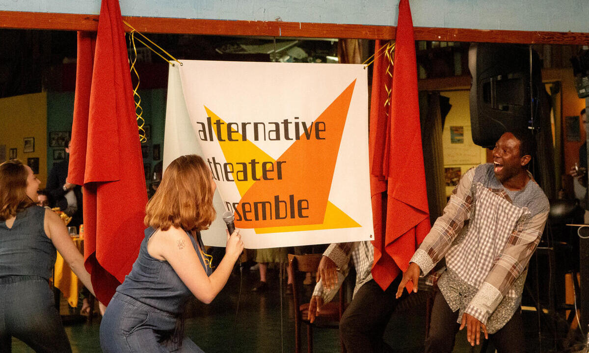 Members of a theater group practicing in front of a "alternative theater ensemble" banner in an informal setting, with a woman speaking into a microphone and a man reacting with laughter.