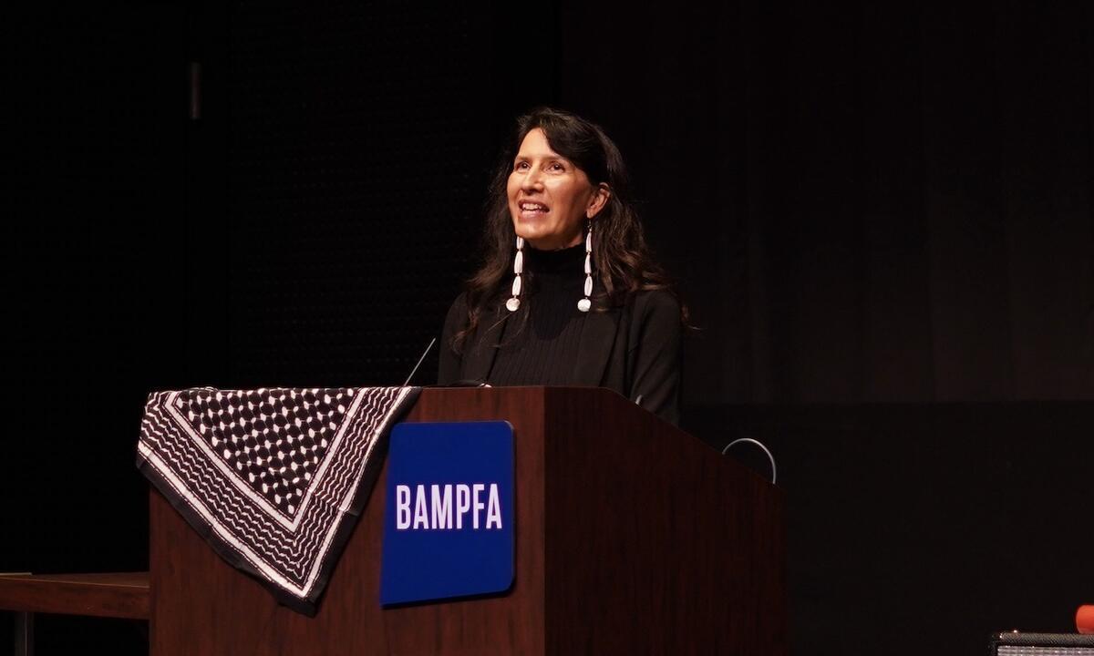 Beth Piatote at a podium. She wears dark clothes and long white earrings.